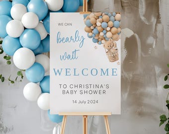 Könnte beinhalten: Ein weißes Willkommens-Schild mit blauem Text, der "We can bearly wait" und "Welcome to Christina's Baby Shower 14 July 2024" lautet. Ein Cartoon-Teddybär in einem Heißluftballon aus blauen und braunen Ballons befindet sich in der oberen rechten Ecke des Schildes.
