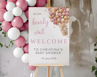 Puede incluir: Un cartel de bienvenida rosa y blanco para un baby shower con el texto "We can bearly wait" y "Welcome to Christina's Baby Shower 14 July 2024". El cartel presenta un oso de peluche de dibujos animados en un globo aerostático con globos rosas y beige.