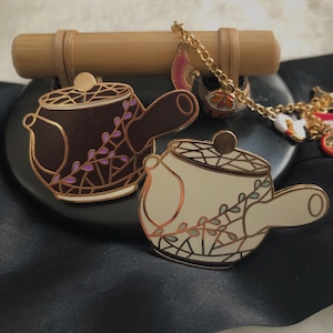 May include: Two decorative teapot pins. One is dark brown with purple accents, the other is off-white with teal accents. Both have gold-colored outlines and lids. A gold chain with charms hangs above the pins, and a bamboo rod rests behind them.