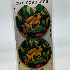 May include: Two round car coasters featuring a colorful frog illustration on a green background with the text "car coasters" and "handmade with love n care".
