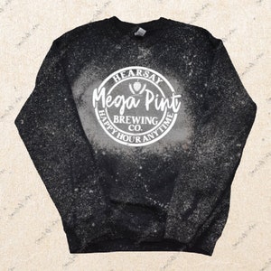 Op de afbeelding: Zwart sweatshirt met een wit ontwerp bespat met bleekmiddel. Het ontwerp bevat een witte cirkel met de tekst "Hearsay Mega Pint Brewing Co. Happy Hour Anytime".