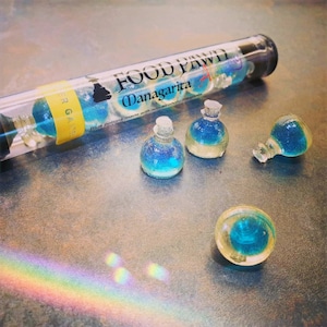 May include: A tube of blue potion bottles labeled "Food Pawd Managarita" with the text "Blue potions made with non-alcoholic margarita mix". The tube is open and there are four blue potion bottles on a gray surface.