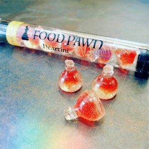 Könnte beinhalten: Drei kleine Glasflaschen gefüllt mit roter und goldener Flüssigkeit. Die Flaschen sind mit "Heartini Alchemy" beschriftet und in einem durchsichtigen Rohr verpackt, auf dem "Food Pawn" steht.