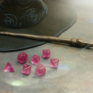 May include: A wooden wand with a carved handle lies on a table next to a black witch's hat and seven pink dice.