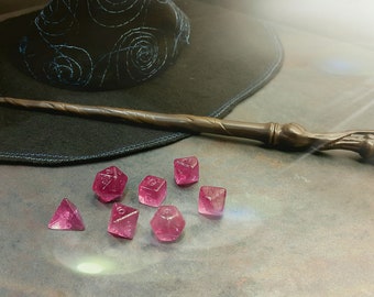 May include: A wooden wand with a carved handle lies on a table next to a black witch's hat and seven pink dice.