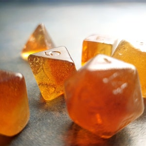 May include: A selection of amber-coloured polyhedral dice. The dice are various shapes, including a four-sided die, a six-sided die, and a twenty-sided die. The dice are made of a clear material with visible inclusions.