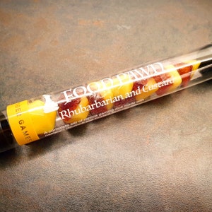 May include: A clear plastic tube filled with yellow and red candy shaped like dice. The tube has a white label that reads "FOOD PAWD Rhubarbarian and Custard A complete set of replica gaming dice made with rhubarb and custard."