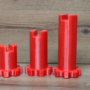 May include: Three red, cylindrical objects with a gear-like base and a notched top are displayed. The objects vary in height, with the tallest one on the right. They are set against a wooden background.