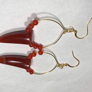 Puede incluir: Un par de pendientes con colgantes en forma de bala de color marr&oacute;n rojizo. Los colgantes est&aacute;n conectados a aros de alambre dorado con peque&ntilde;as cuentas redondas de color naranja. Los pendientes tienen un cierre de gancho.