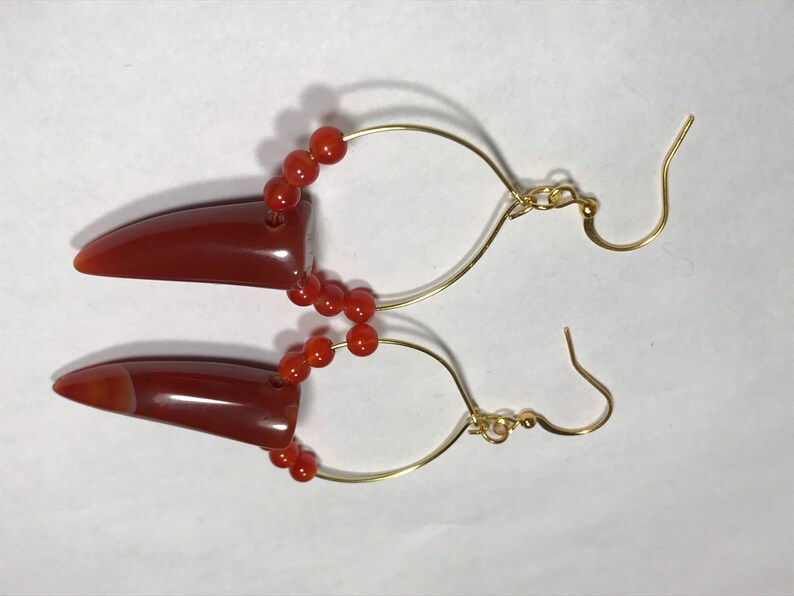 Puede incluir: Un par de pendientes con colgantes en forma de bala de color marr&oacute;n rojizo. Los colgantes est&aacute;n conectados a aros de alambre dorado con peque&ntilde;as cuentas redondas de color naranja. Los pendientes tienen un cierre de gancho.