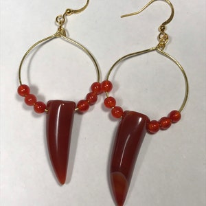 Puede incluir: Un par de pendientes de aro dorados. Cada pendiente presenta un colgante pulido en forma de diente, de color marr&oacute;n rojizo, suspendido de un aro dorado. Peque&ntilde;as cuentas redondas de color naranja est&aacute;n ensartadas en la parte inferior de los aros.