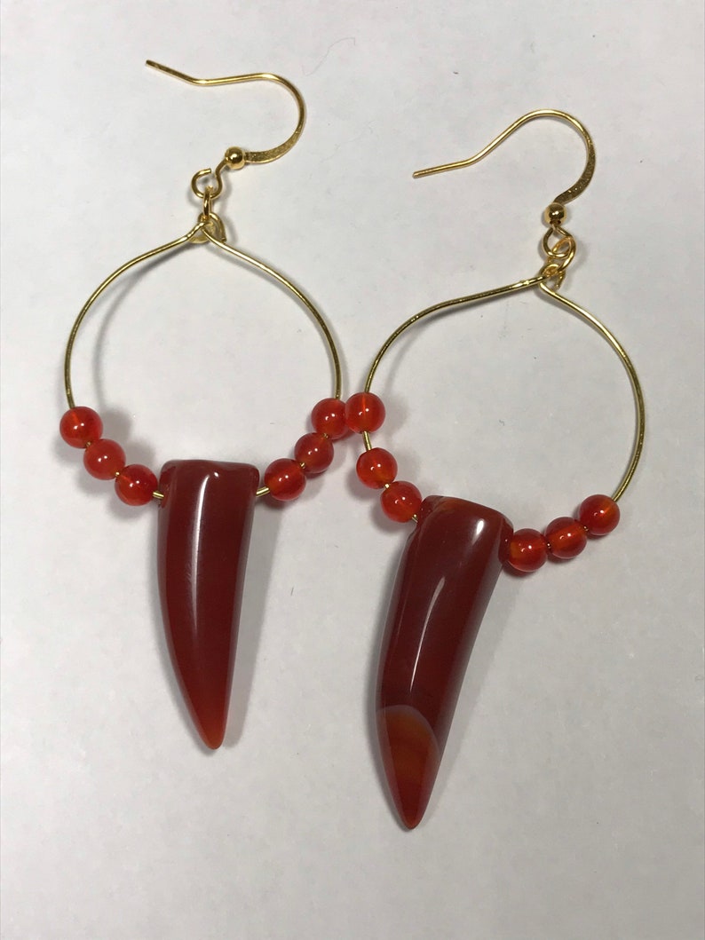 Puede incluir: Un par de pendientes de aro dorados. Cada pendiente presenta un colgante pulido en forma de diente, de color marr&oacute;n rojizo, suspendido de un aro dorado. Peque&ntilde;as cuentas redondas de color naranja est&aacute;n ensartadas en la parte inferior de los aros.