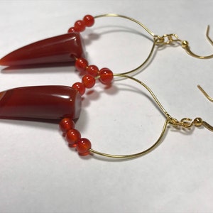 Puede incluir: Un par de pendientes hechos a mano con colgantes de &aacute;gata pulida de color marr&oacute;n rojizo con forma puntiaguda. Los pendientes tienen ganchos dorados curvados y est&aacute;n adornados con peque&ntilde;as cuentas redondas de color naranja. El dise&ntilde;o es sencillo y elegante.