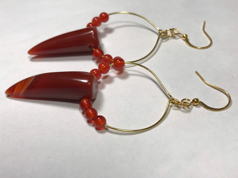 Puede incluir: Un par de pendientes hechos a mano con colgantes de &aacute;gata pulida de color marr&oacute;n rojizo con forma puntiaguda. Los pendientes tienen ganchos dorados curvados y est&aacute;n adornados con peque&ntilde;as cuentas redondas de color naranja. El dise&ntilde;o es sencillo y elegante.