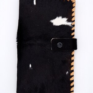 Puede incluir: Cartera de piel de vaca negra y blanca con un borde cosido marr&oacute;n y una solapa de cuero negro con cierre de presi&oacute;n.