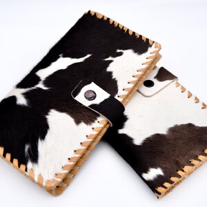 Puede incluir: Dos carteras de piel de vaca en blanco y negro con costuras marrones y una correa de cuero negro en una de las carteras.