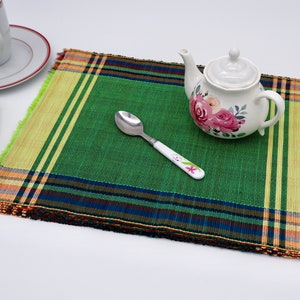 May include: A green placemat with a yellow, orange, red, blue and black plaid border. A white teapot with pink roses and green leaves is on the placemat, along with a white spoon with pink flowers.