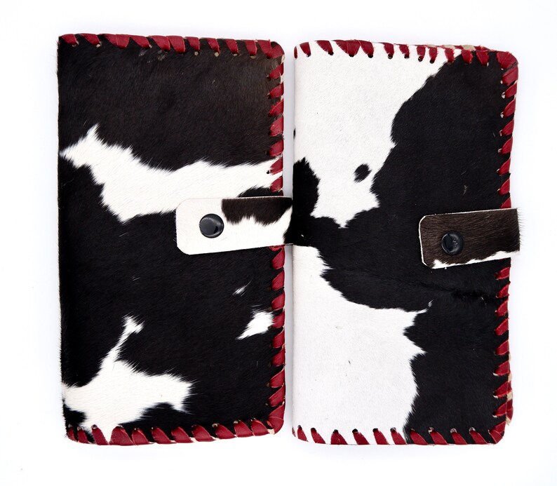 Puede incluir: Dos carteras de piel de vaca negra y blanca con costuras rojas. Las carteras tienen un cierre de solapa con un bot&oacute;n.