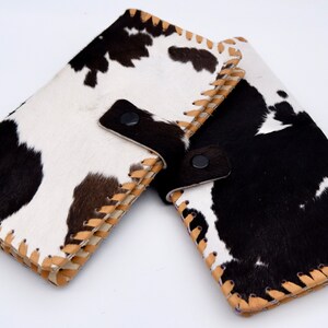 Puede incluir: Dos carteras de piel de vaca en blanco y negro con costuras marrones y un cierre de presi&oacute;n negro.