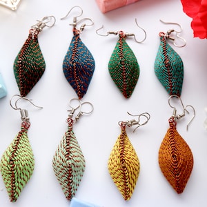 May include: Eight pairs of handmade earrings with a diamond shape. The earrings are made of woven fabric in various colours, including green, blue, yellow, and orange. Each earring has a silver hook.