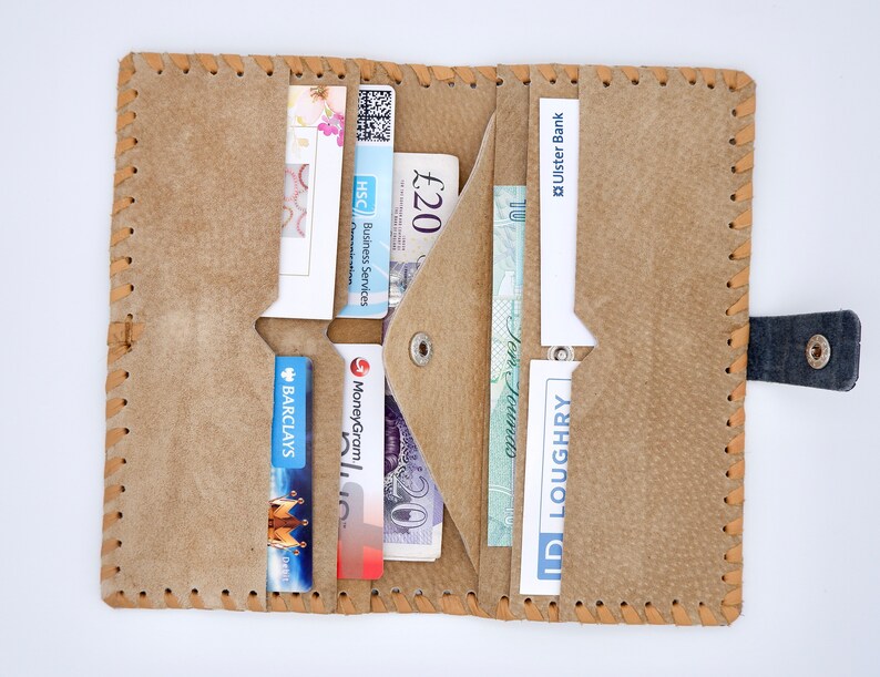 Puede incluir: Una cartera de cuero marr&oacute;n con m&uacute;ltiples ranuras para tarjetas y una solapa para efectivo. La cartera est&aacute; abierta y muestra varias tarjetas, incluida una tarjeta de Barclay's, una tarjeta de MoneyGram y una tarjeta de HSC Business Services. Tambi&eacute;n hay varios billetes de libra esterlina dentro de la cartera.