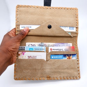 Puede incluir: Una cartera de cuero marr&oacute;n con m&uacute;ltiples ranuras para tarjetas y un portafolios. La cartera est&aacute; abierta y muestra varias tarjetas y billetes en el interior, incluyendo una tarjeta MoneyGram, una tarjeta Barclays y un billete de 20 libras.