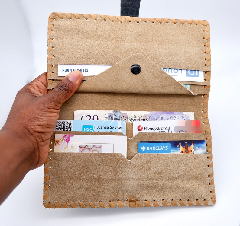 Puede incluir: Una cartera de cuero marr&oacute;n con m&uacute;ltiples ranuras para tarjetas y un portafolios. La cartera est&aacute; abierta y muestra varias tarjetas y billetes en el interior, incluyendo una tarjeta MoneyGram, una tarjeta Barclays y un billete de 20 libras.