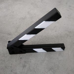 May include: A black and white film clapperboard, open and angled. The clapperboard has alternating black and white stripes on the top and bottom sections. The clapperboard is on a grey surface.