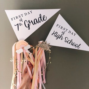 May include: Two white triangular pennants with black text. The top pennant reads "First Day of 7th Grade". The bottom pennant reads "First Day of High School". A hand holds a gold headband with pink and yellow ribbons.