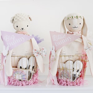 May include: Two pastel pink bunny baskets filled with pink and white shredded paper. One basket has a white bunny doll with a pink bow and a sign that says "Big Bunny". The other basket has a white bunny doll with a flower crown and a sign that says "Little Bunny".