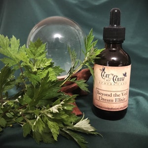 May include: A brown glass bottle with a dropper top and a label that reads "Cat & Crow Apothecary Beyond the Veil Dream Elixir catandcrowapothecary.etsy.com". The bottle is sitting on a green surface next to a clear glass orb.