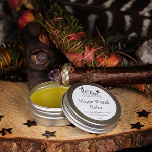 May include: A small silver tin with a yellow balm inside. The lid of the tin is open and shows a label that reads "Cat & Crow Apothecary Magic Wand Balm Calendula, Plantain, Yarrow Olive Oil, Beeswax 1 oz."