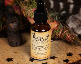 May include: A brown glass bottle with a dropper top and a label that reads "Cat & Crow Apothecary Skullcap Tincture Scutellaria Lateriflora 100 Proof Vodka etsy.com/shop/catandcrowapothecary".