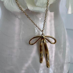 May include: A gold-colored necklace with a bow pendant. The bow pendant has two elongated, curved sections hanging below two loops. The chain is a delicate, linked design. The necklace is displayed against a white background.