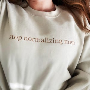 Puede incluir: Sudadera verde claro con la frase "stop normalizing men" impresa en letras marrones. La sudadera de cuello redondo es de color liso y tiene un corte holgado.