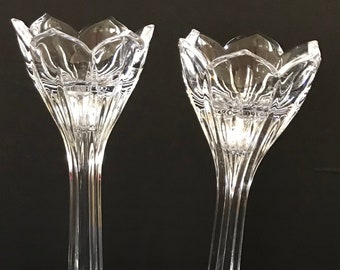 May include: A pair of clear glass candle holders with a tulip-shaped design. The holders have a tall, slender stem and a wide, flared base.