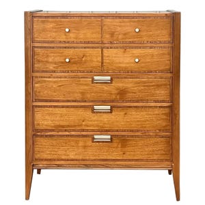 May include: A six-drawer dresser made of wood with a light brown finish. The drawers have silver metal pulls and are accented with a thin, dark brown line around the edges.