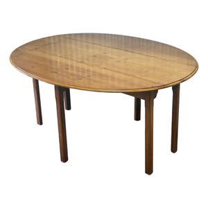 May include: An oval wooden dining table with a smooth, light brown finish showing natural wood grain patterns. The table features a drop-leaf design with four straight, sturdy legs that provide support for the large, rounded tabletop surface.