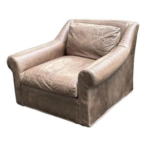 May include: A light brown leather armchair with a matching pillow. The chair has rolled arms and a low back. The leather appears worn, with visible creases and variations in color, giving it a vintage look. The chair is designed for comfort and relaxation.