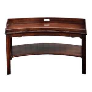 May include: A dark brown wooden serving table with a curved top and a lower shelf. The top has a handle cut-out. The table has four legs and is likely made of solid wood. The style is likely antique or vintage.