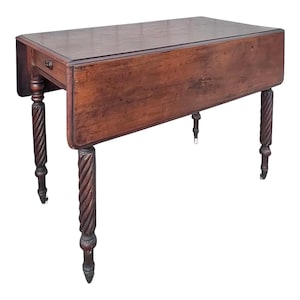 May include: A rectangular brown wooden drop-leaf table featuring a smooth tabletop surface and four legs with a distinct spiral-turned design. The table includes a small drawer with a round knob and is supported by metal casters on each leg.