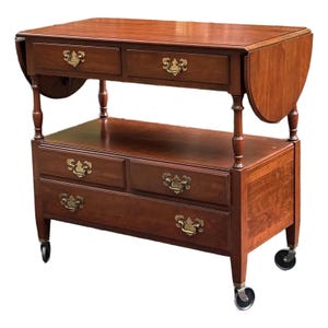 May include: A cherry-wood tea cart with two drop leaves, two drawers on the top level, and two drawers on the bottom level. The cart has brass hardware and sits on four caster wheels.