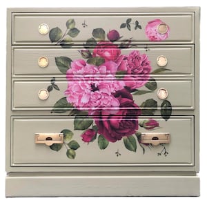 May include: A sage green chest of drawers with four drawers. The drawers are adorned with a large floral decal featuring pink and burgundy peonies and roses, with green leaves. The hardware is gold-toned.