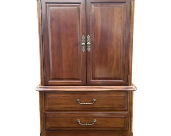 May include: A tall, wooden armoire with a warm brown finish. The upper section features two doors with ornate brass handles. Below, two drawers with matching handles provide additional storage. The armoire has a classic, traditional design.