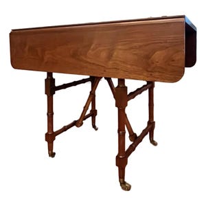 May include: A brown wooden drop-leaf table with a rectangular top and a smooth grain finish. The table features four legs designed with a bamboo-style texture, each ending in a small brass caster wheel for mobility, supporting the folding side leaves.