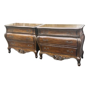 May include: Two matching antique wooden dressers with three drawers each. The dressers have a curved front design, ornate hardware, and decorative carvings. The wood has a warm, brown finish, and the dressers are set against a white background.