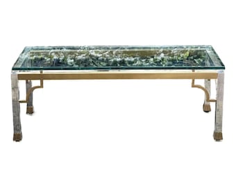 May include: A rectangular coffee table with a glass top and a decorative interior filled with greenery. The table has a gold-colored frame and silver-colored legs. The table is approximately 48 inches long and 24 inches wide.