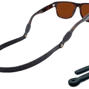 K&ouml;nnte beinhalten: Ein schwarzes Leder-Sonnenbrillenband mit goldenen Beschl&auml;gen und zwei schwarzen Gummib&auml;ndern f&uuml;r Sonnenbrillen. Das Band ist an einer Sonnenbrille aus Schildpatt mit braunen Gl&auml;sern befestigt.