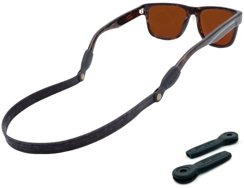 K&ouml;nnte beinhalten: Ein schwarzes Leder-Sonnenbrillenband mit goldenen Beschl&auml;gen und zwei schwarzen Gummib&auml;ndern f&uuml;r Sonnenbrillen. Das Band ist an einer Sonnenbrille aus Schildpatt mit braunen Gl&auml;sern befestigt.