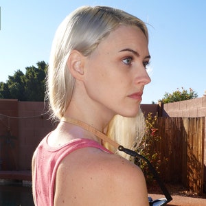 K&ouml;nnte beinhalten: Eine Frau mit blonden Haaren tr&auml;gt ein rosafarbenes Tanktop und eine braune Lederkameragurt. Der Gurt ist an einem schwarzen Kameraobjektiv befestigt.